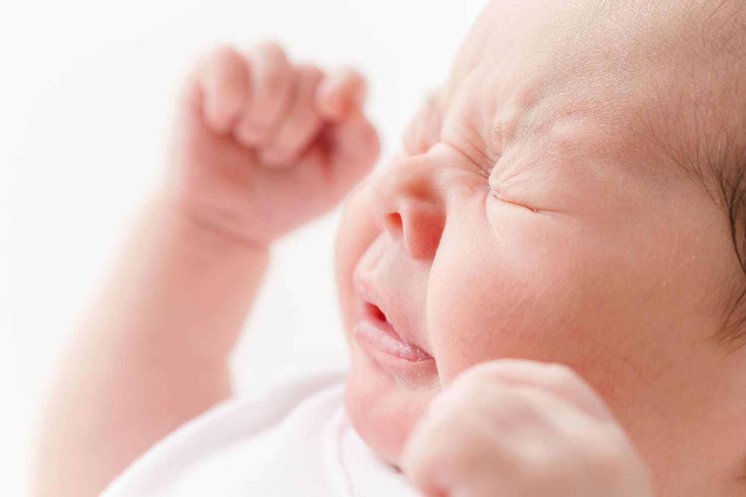 Close-up of a newborn baby yawning or crying with tiny fist raised against soft pink background.