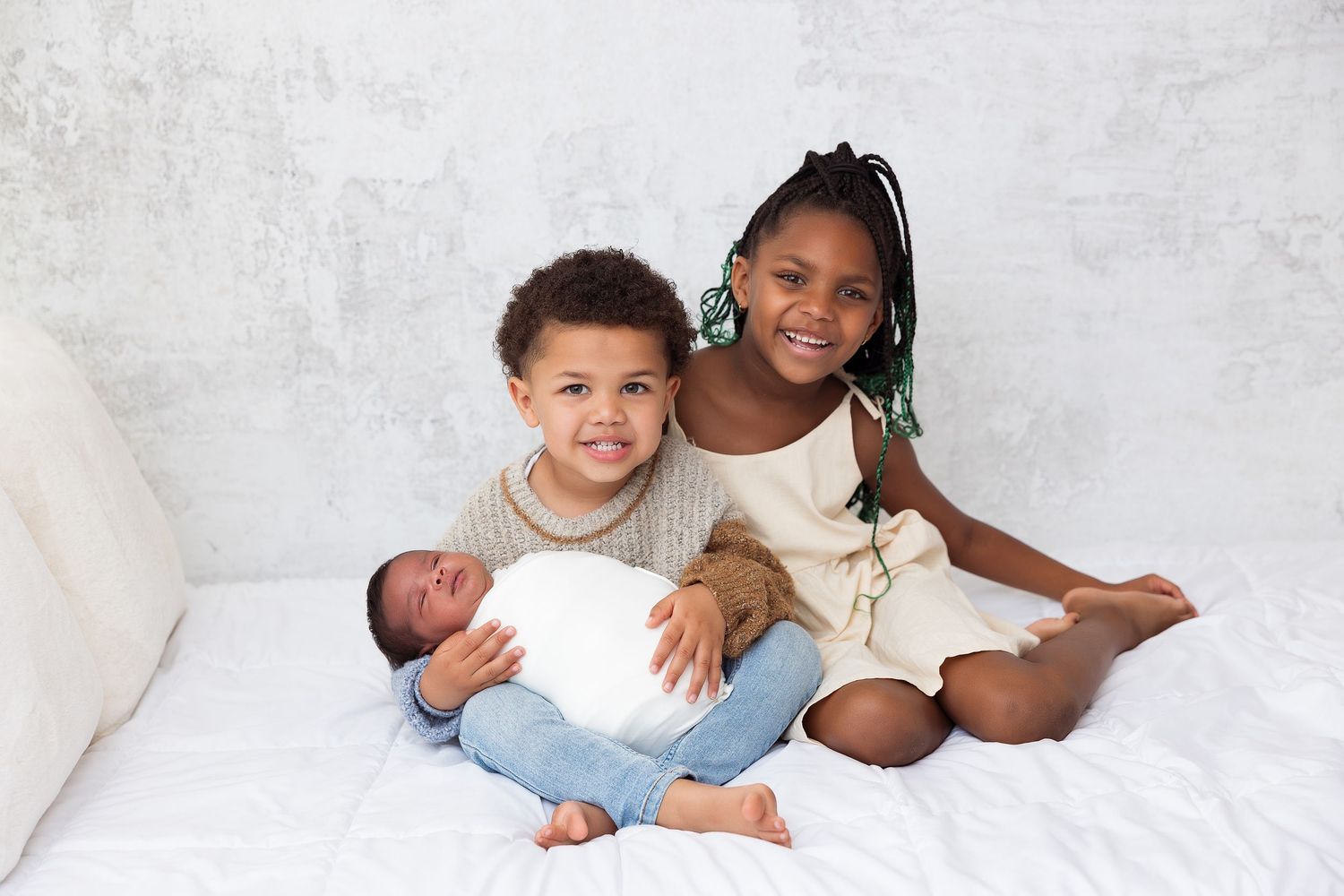 Children sitting on white bedding with a newborn baby wrapped in blue blanket.