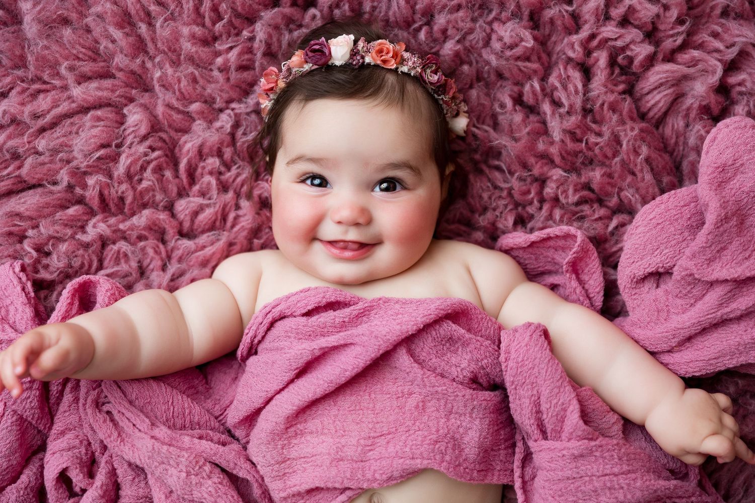 Baby wearing floral headband lies on plush pink blanket during photo session.