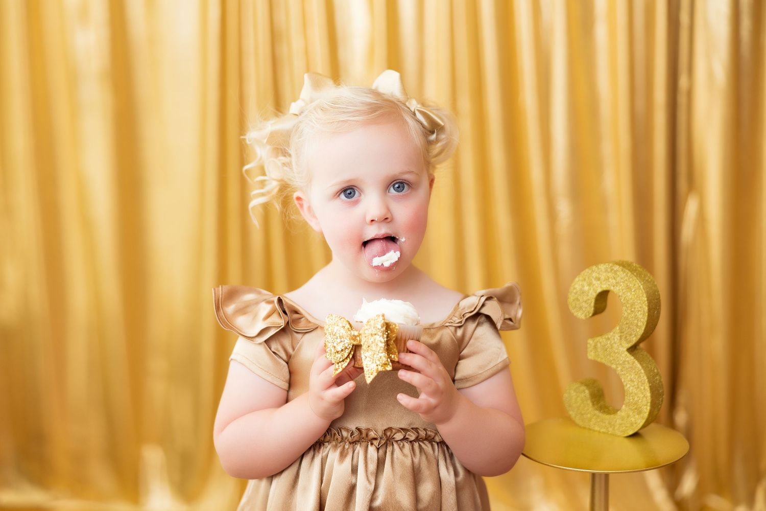 Child in gold dress poses with number 3 decoration against gold curtain backdrop while holding celebratory treat.
