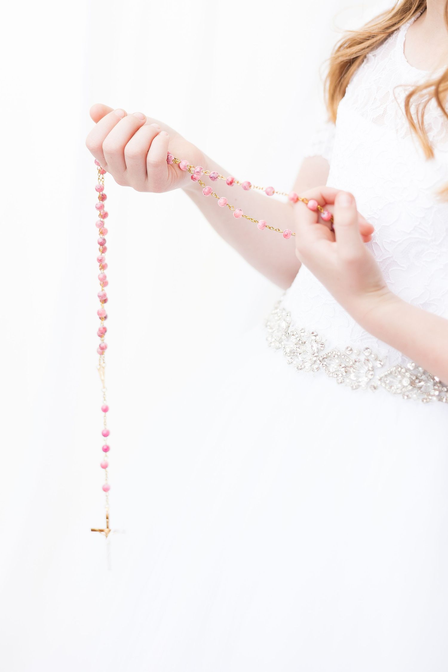 Hands in white lace sleeves holding a pink rosary against a bright background.