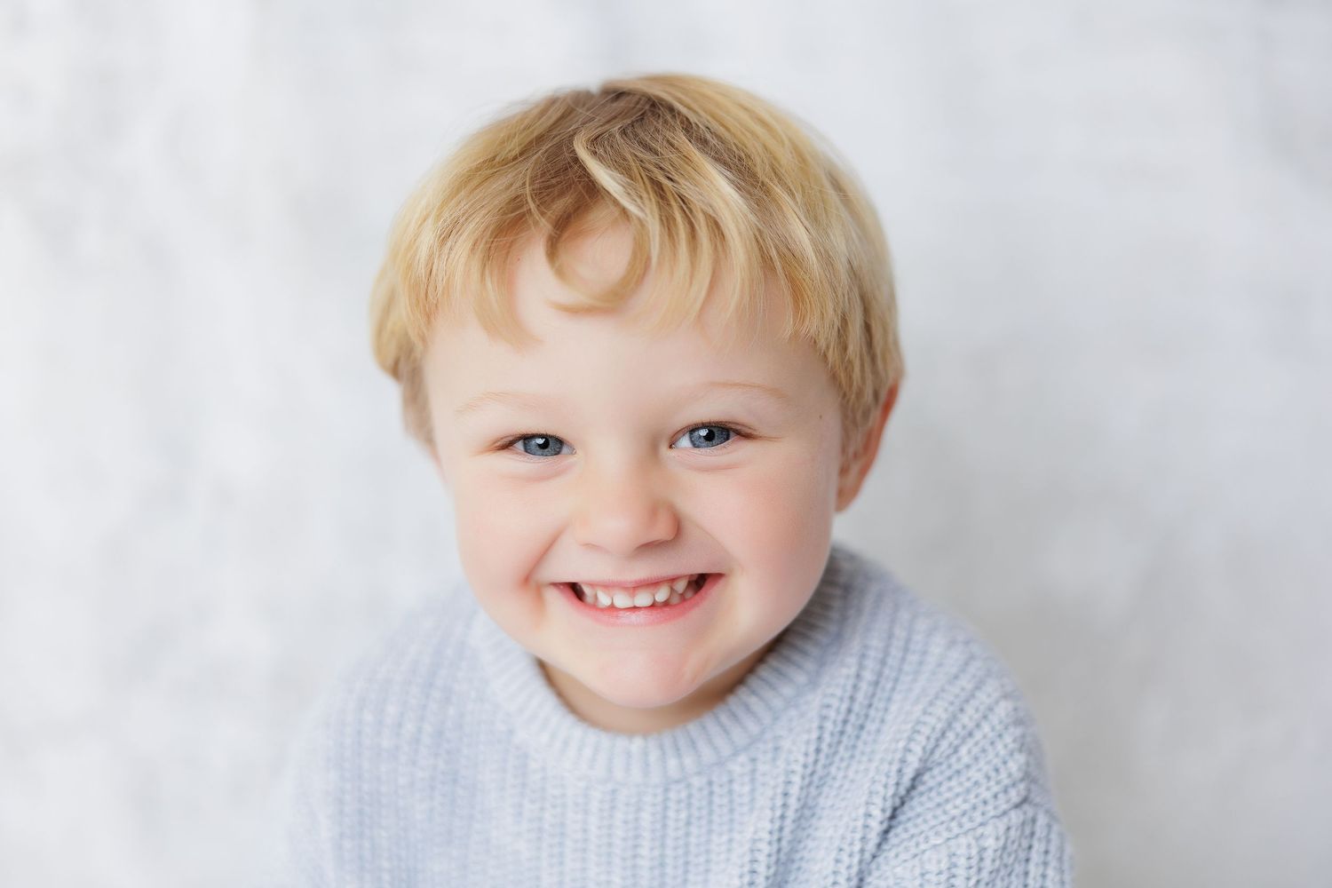 Smiling toddler boy with blonde hair and blue eyes wearing a light blue knit sweater against white background.