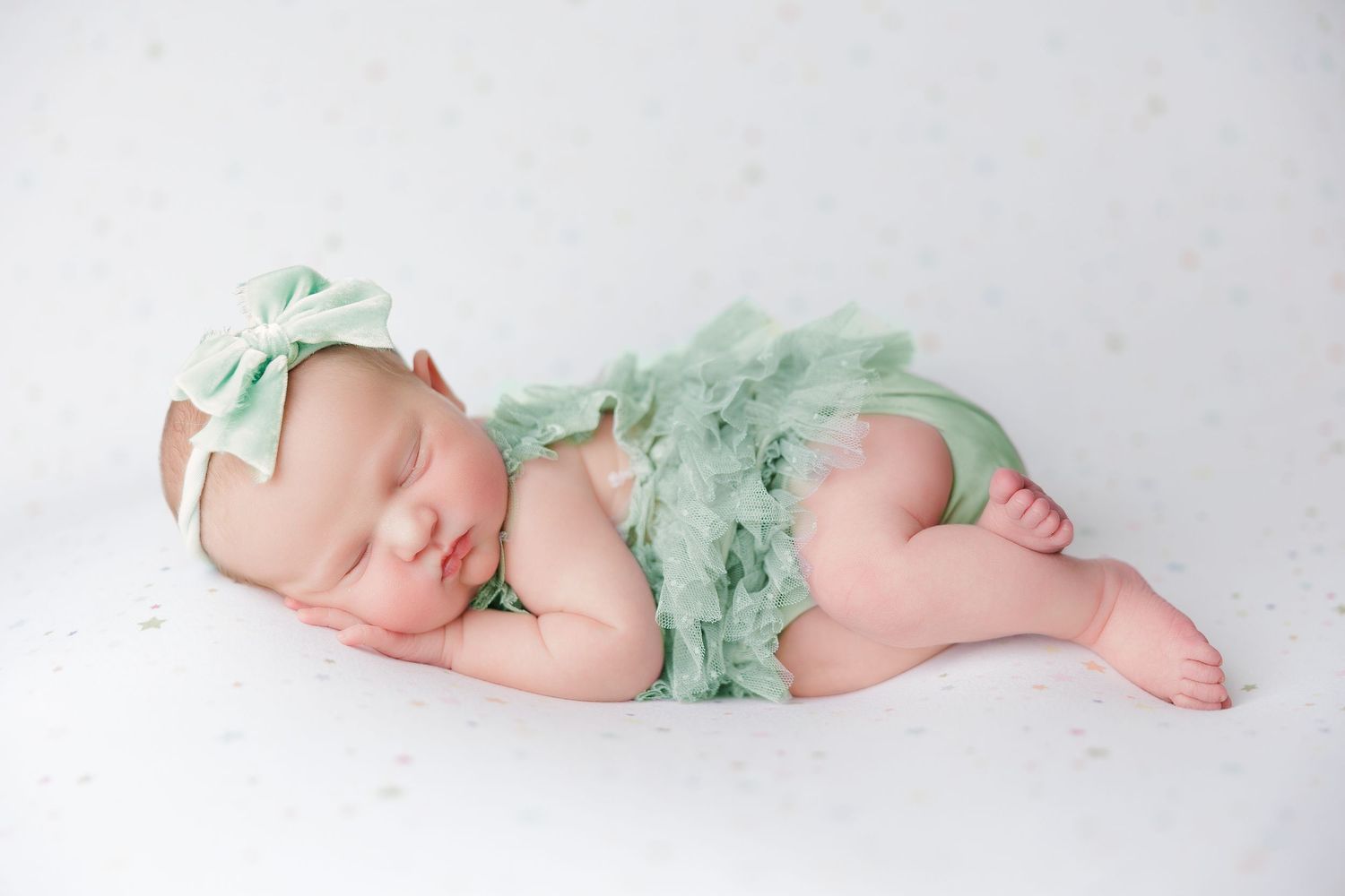 Newborn baby girl sleeping peacefully in a mint green ruffled outfit and bow headband on a white background.