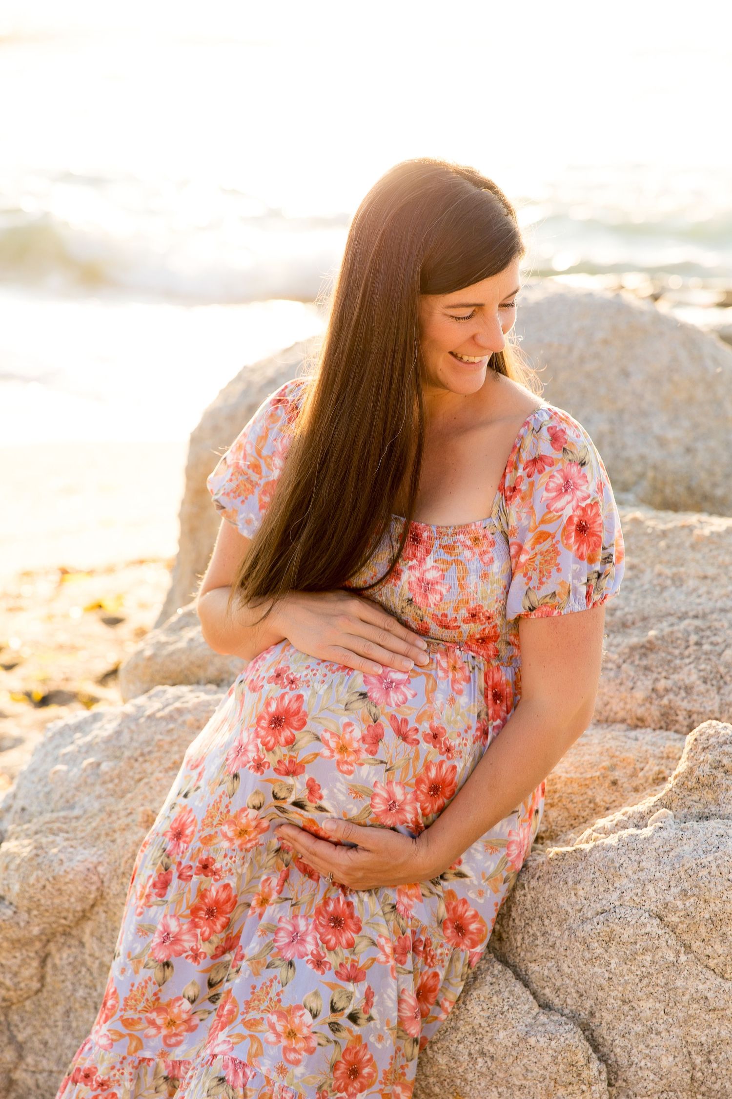 Expectant mother in floral dress sits on coastal rocks during golden hour photography session.