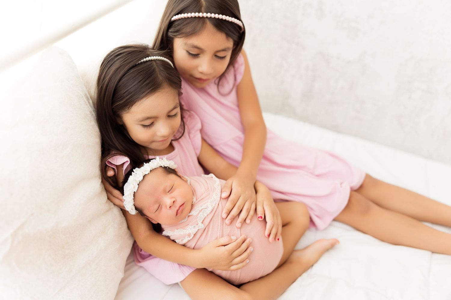 Sisters in pink dresses cuddling together with infant on white background.