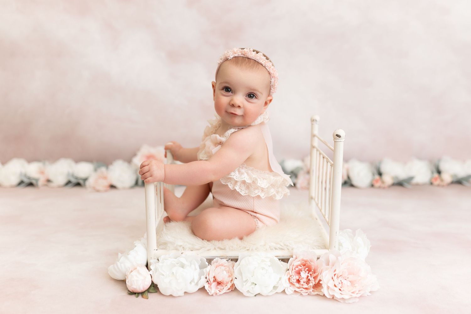 Baby seated by vintage bed with pink and white floral decorations.