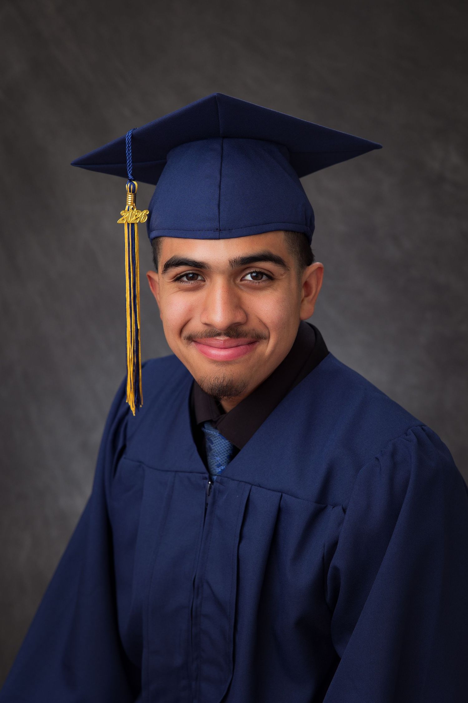 Young man smiling in navy blue graduation cap and gown with gold tassel, posing for formal graduation portrait.