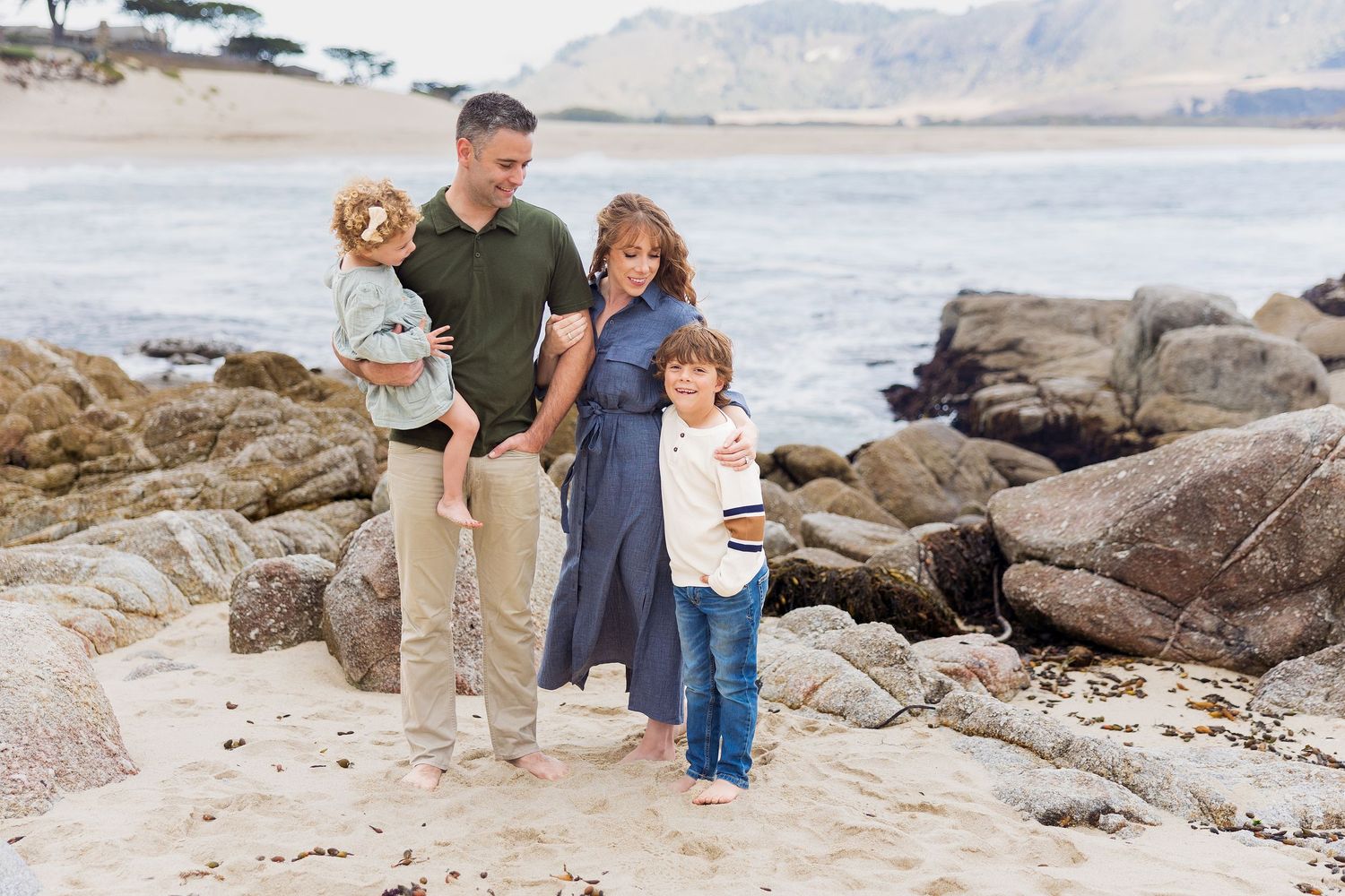 Family stands together on rocky beach with ocean waves and mountains visible behind them.