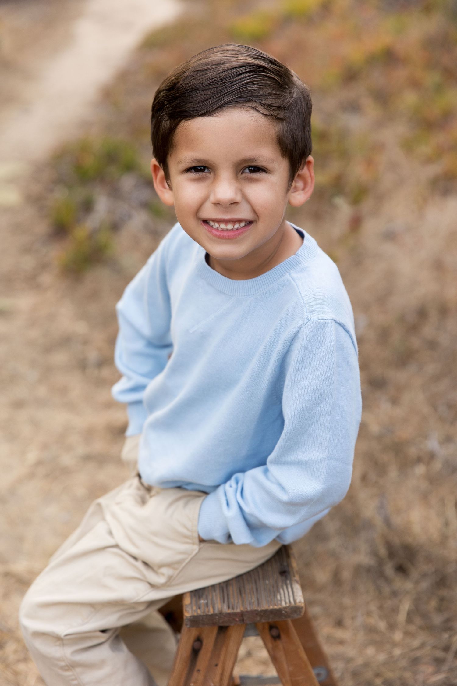 Child in light blue sweater sits casually on wooden stool for outdoor portrait.