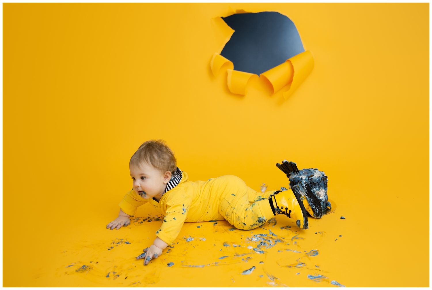 Baby in graduation cap crawls through colorful confetti on bright yellow background creating playful celebration scene.