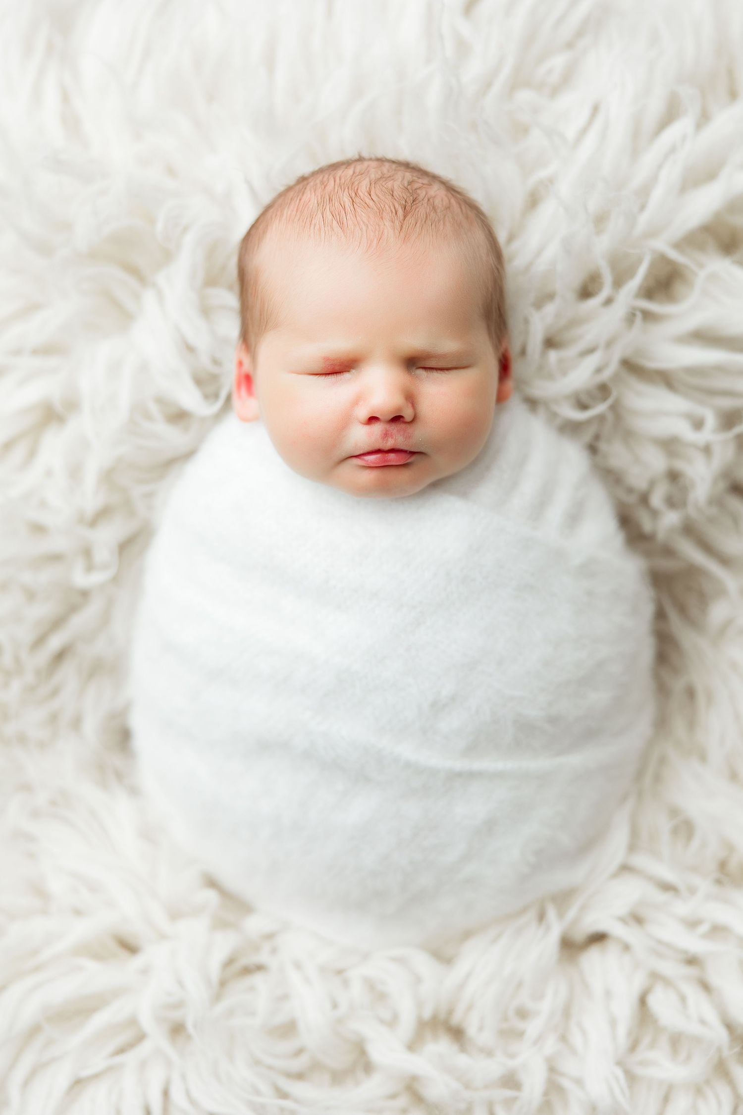 Peaceful sleeping newborn baby swaddled in white blanket resting on soft white fur background.