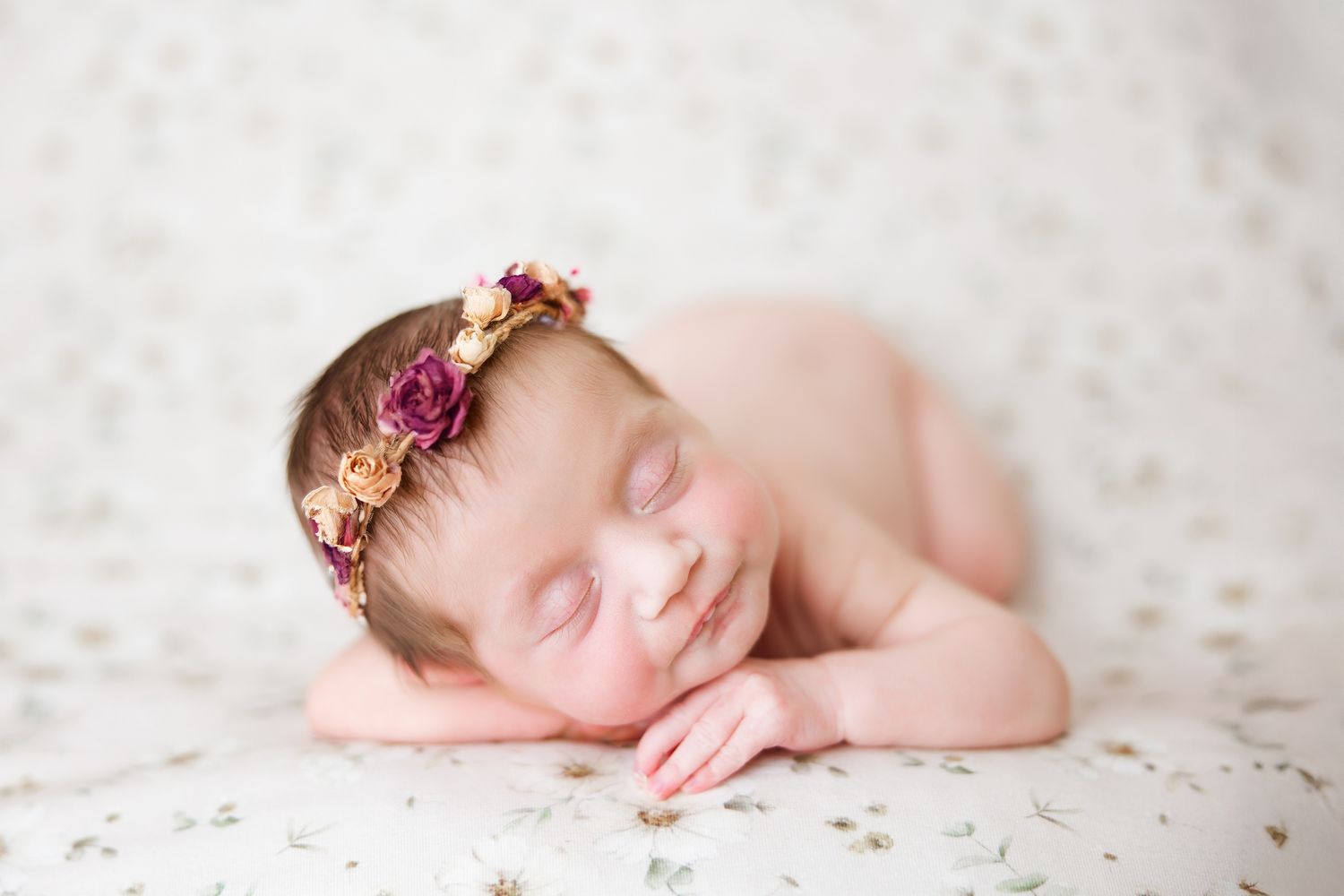 Sleeping newborn baby wearing delicate pink floral crown rests peacefully on white textured blanket.
