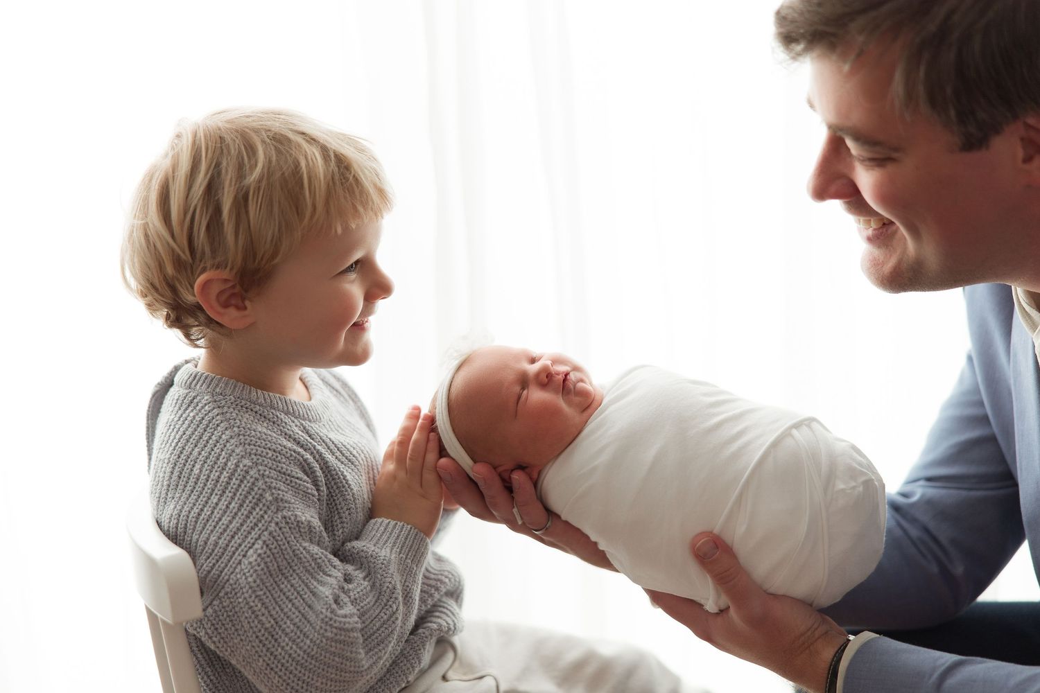 Father holds newborn baby while young toddler sibling smiles and reaches out to touch the infant.