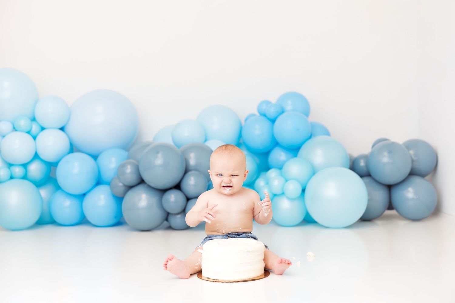 Baby in diapers sits beside decorative blue and gray balloon garland backdrop for cake smash photoshoot.