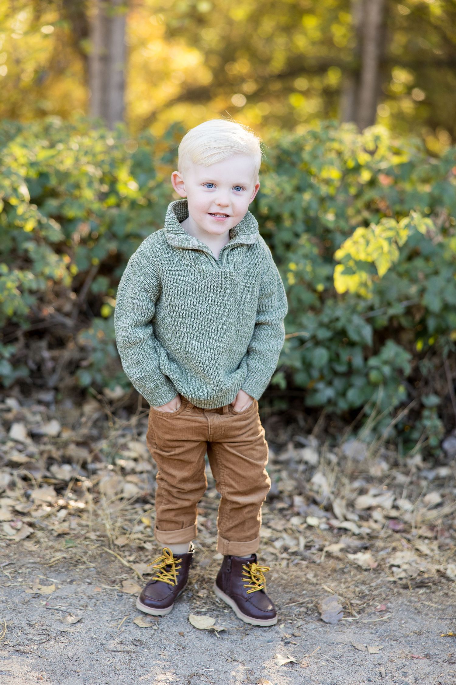 Small child in a gray sweater and brown pants standing on fallen leaves.