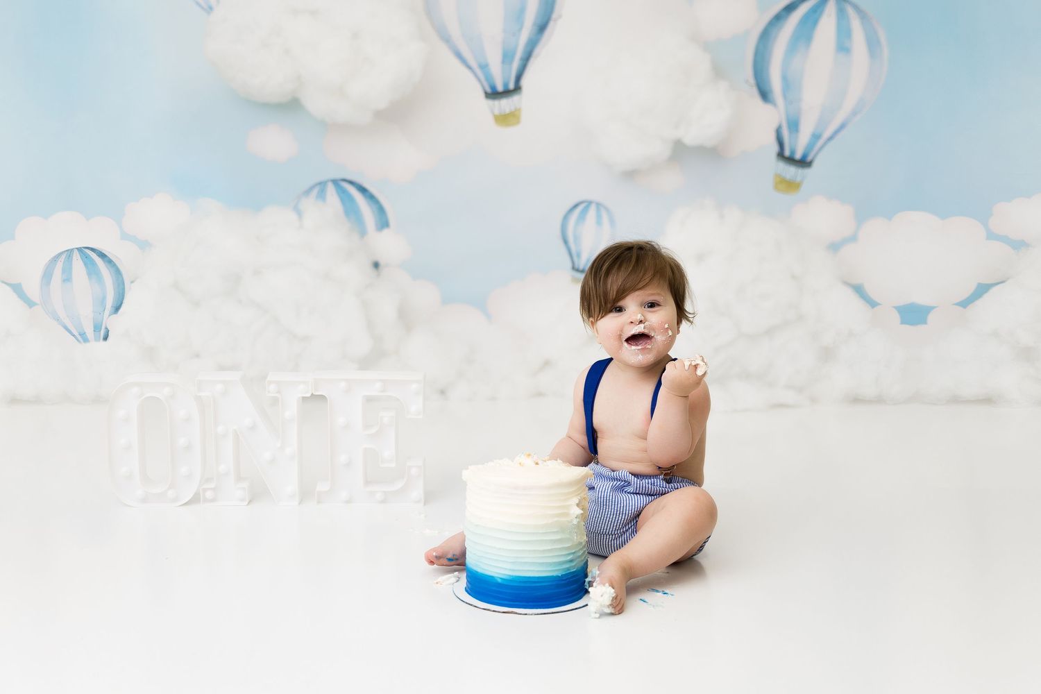 Baby sitting on white floor with blue hot air balloon backdrop and birthday cake smash setup.