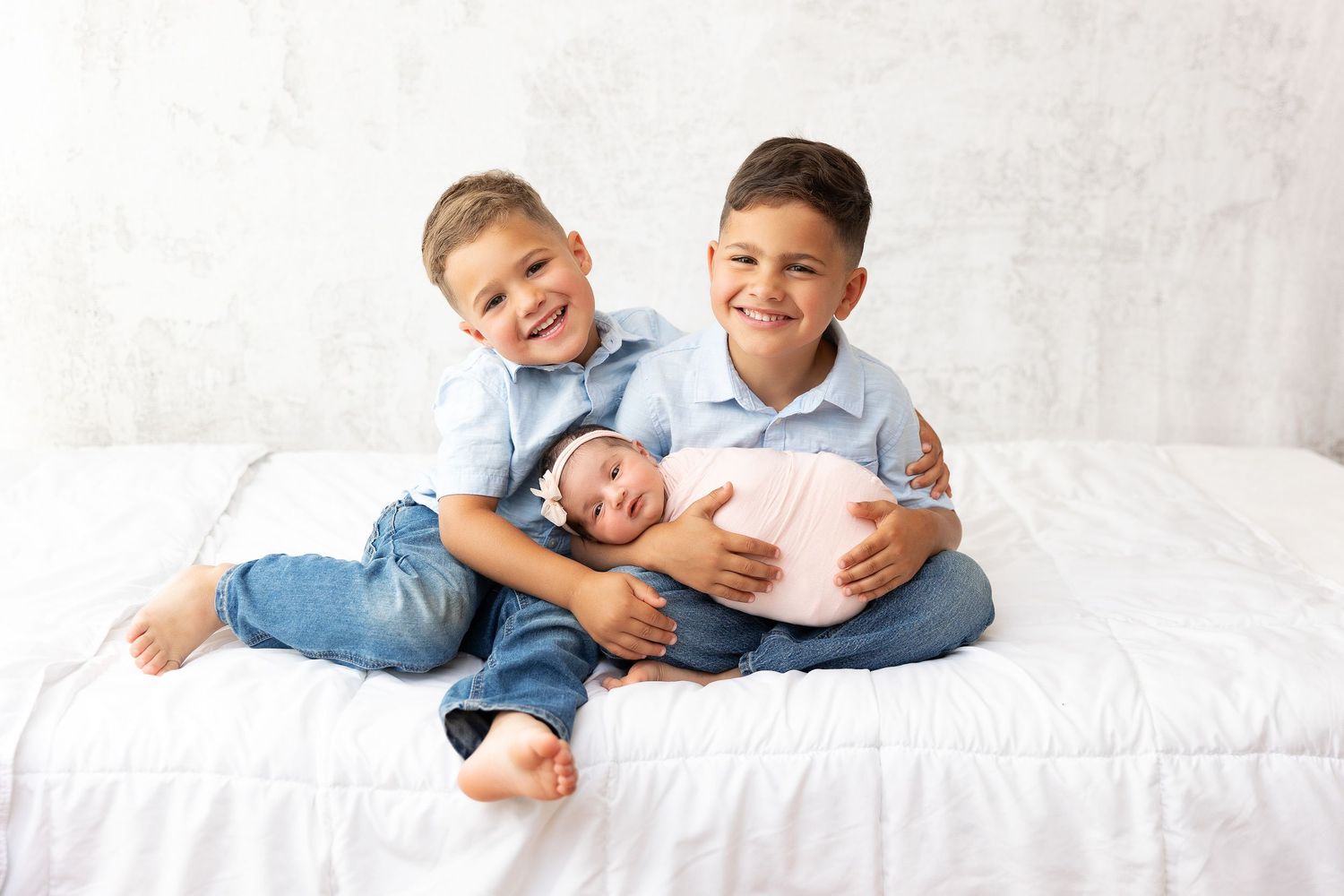 Three children in light blue shirts sitting together on white bed with newborn sibling.