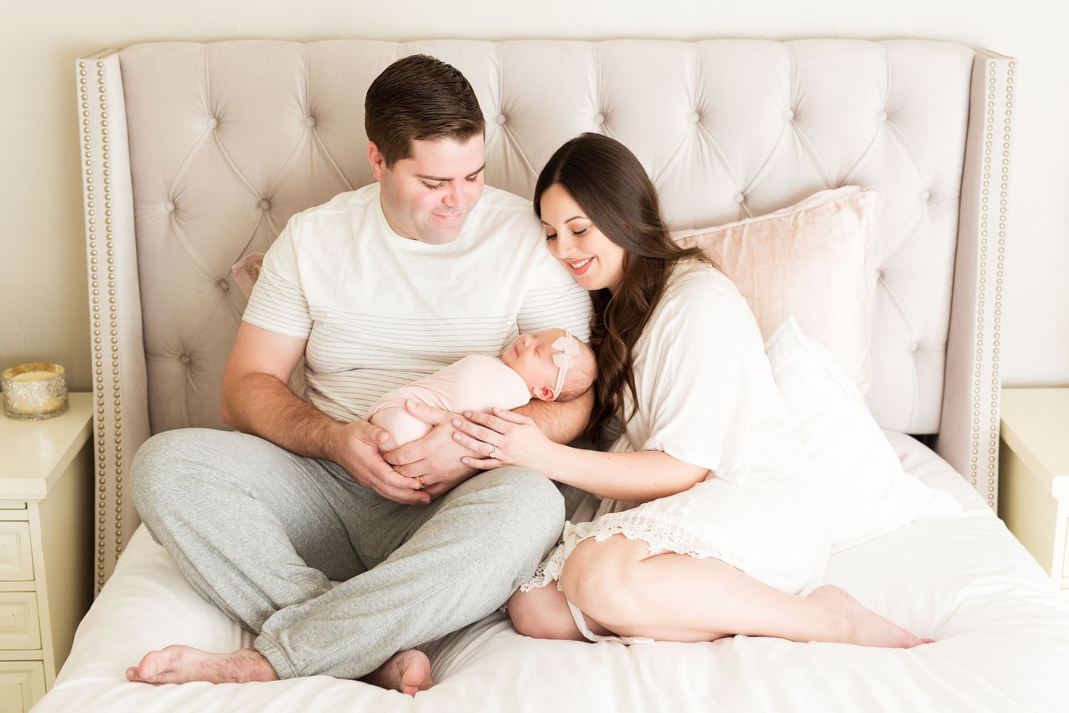Parents lovingly cradling newborn baby while sitting on beige tufted headboard bed.