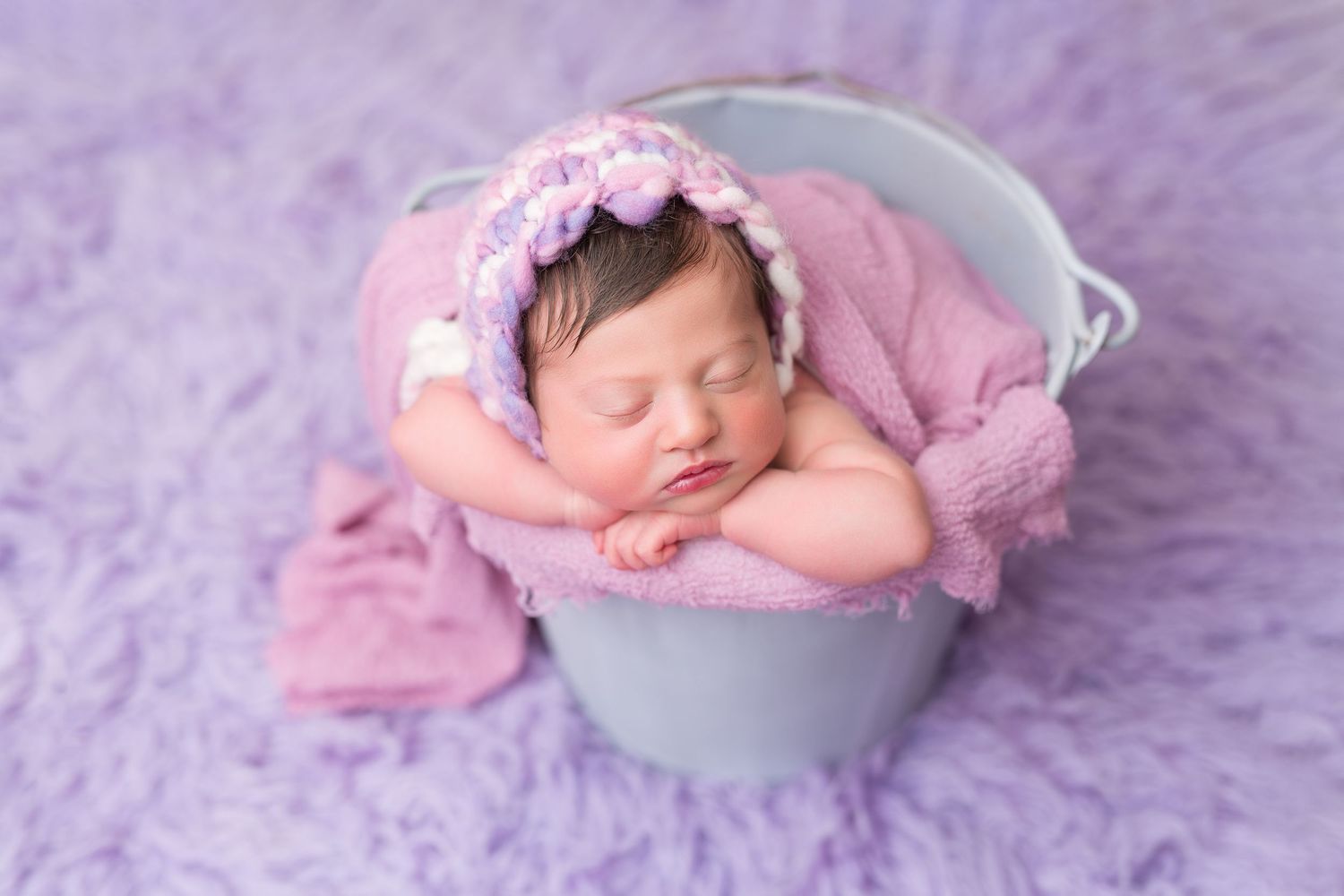 Sleeping newborn in purple bonnet posed in gray bucket with pink fabric.