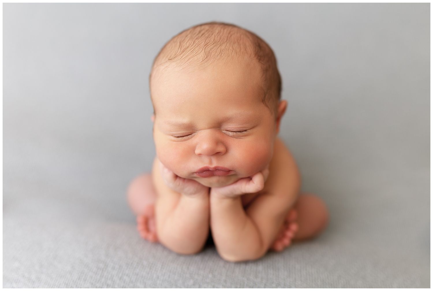 Peaceful sleeping newborn baby posed with hands under chin on soft gray backdrop.