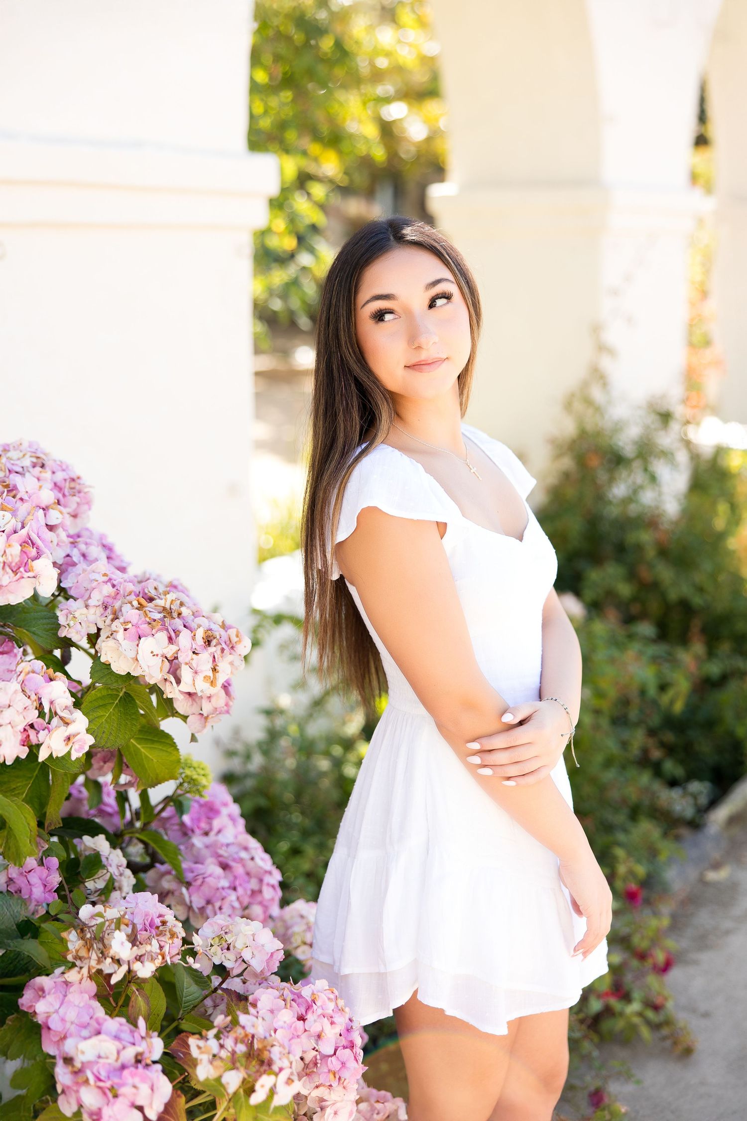 Young woman in white mini dress standing beside pink hydrangeas in sunny garden.