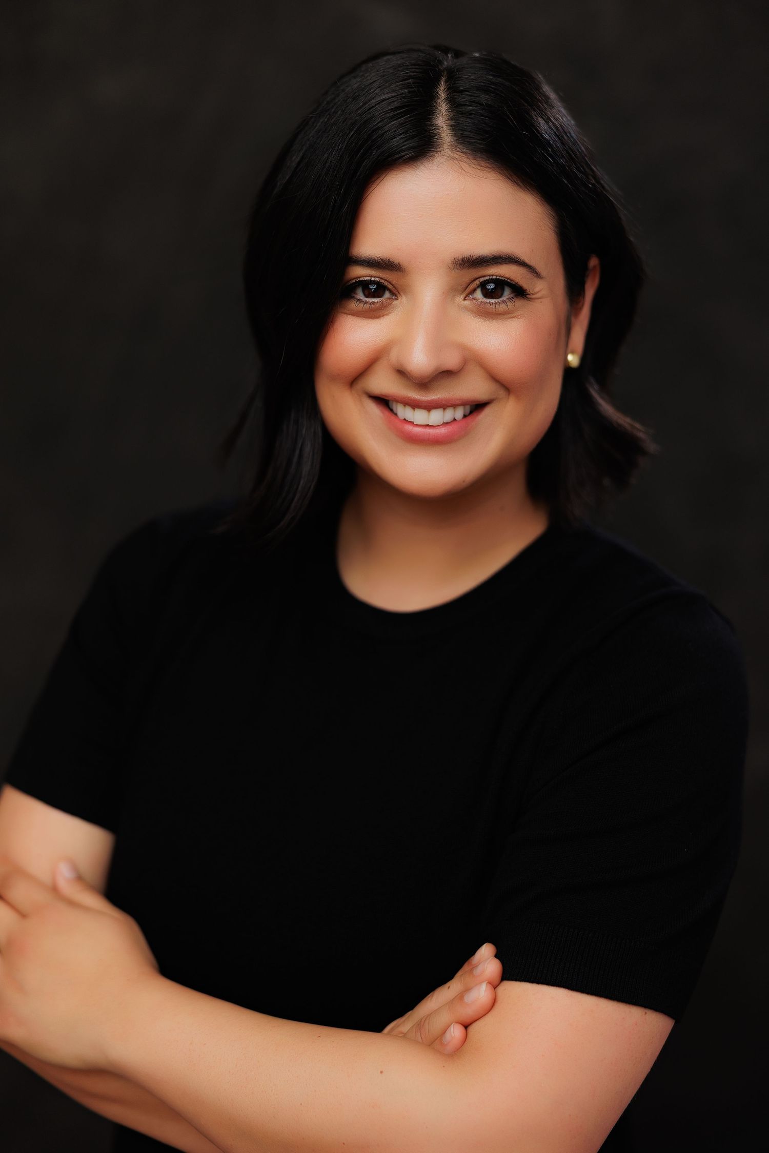 Smiling woman with dark hair in black t-shirt, arms crossed, professional headshot on dark background.