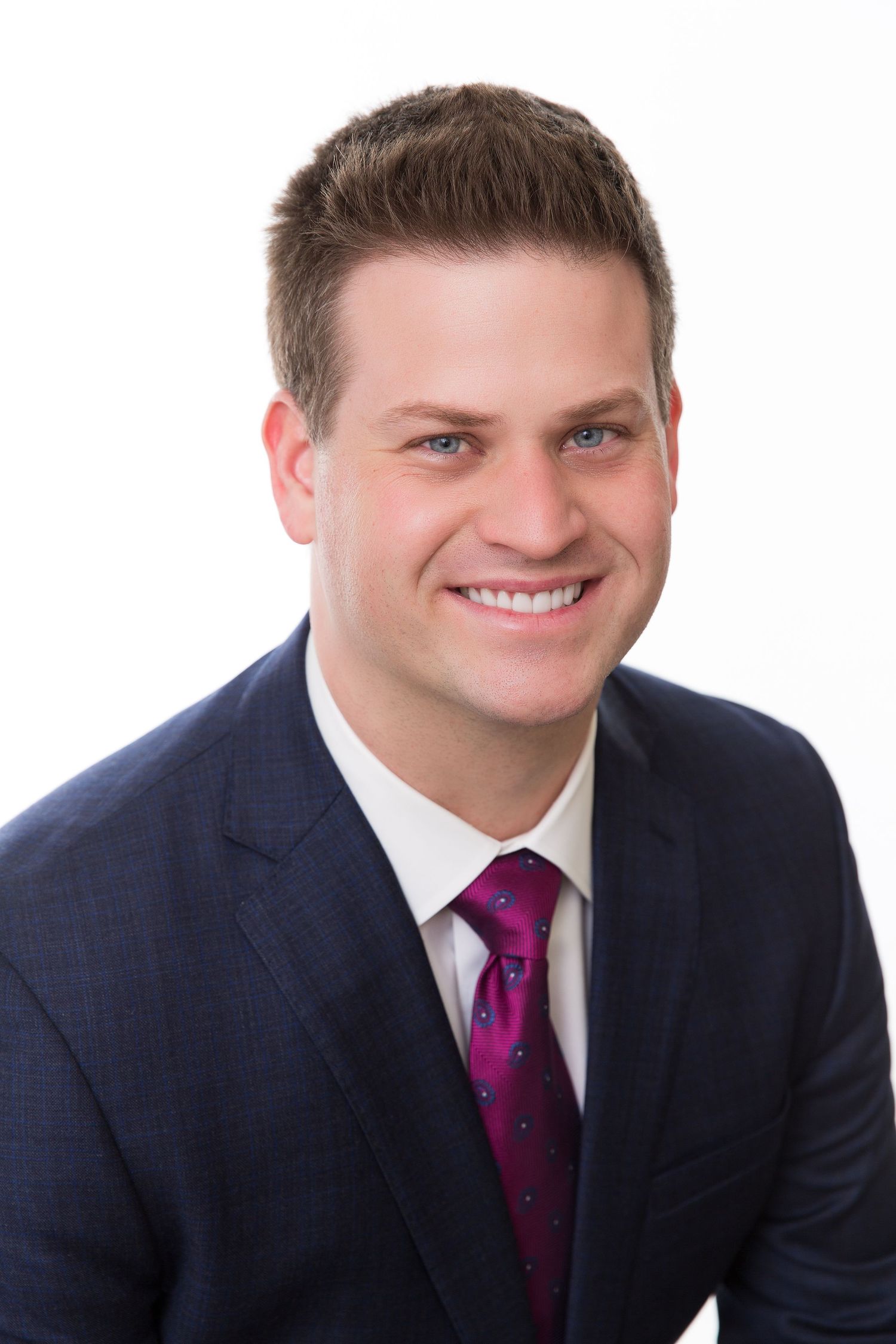 Professional headshot of a smiling young man in a navy suit and magenta tie against a white background.