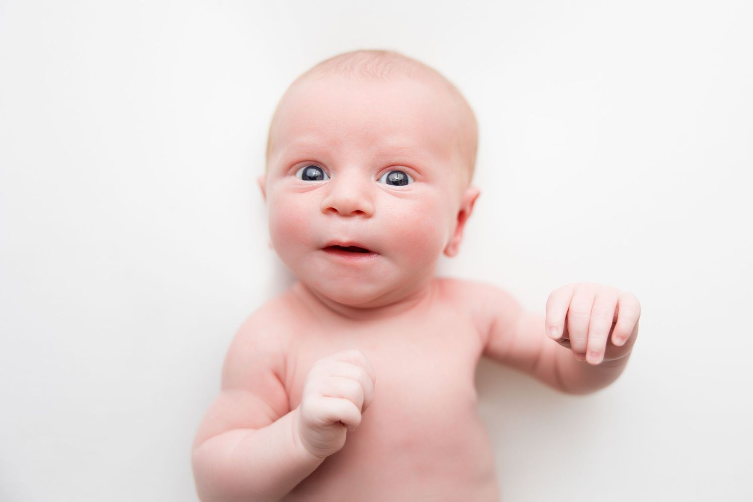 A baby lying on white fabric with a curious expression and reaching out with one hand.