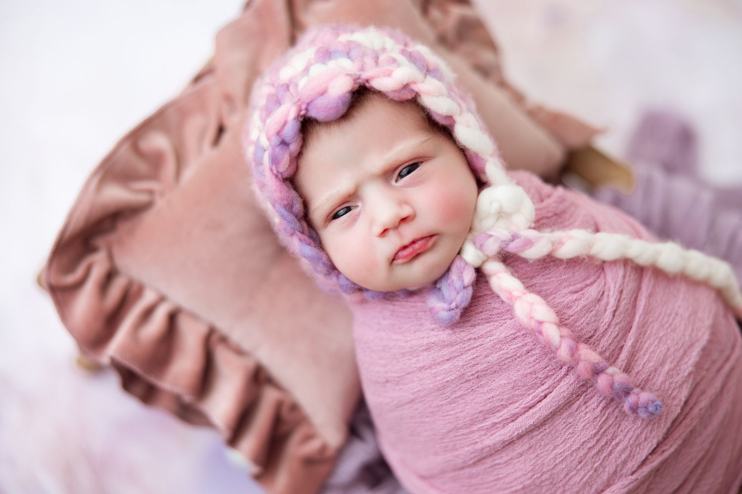 Newborn baby wrapped in soft pink swaddle blanket with matching headband lying peacefully on ruffled fabric.