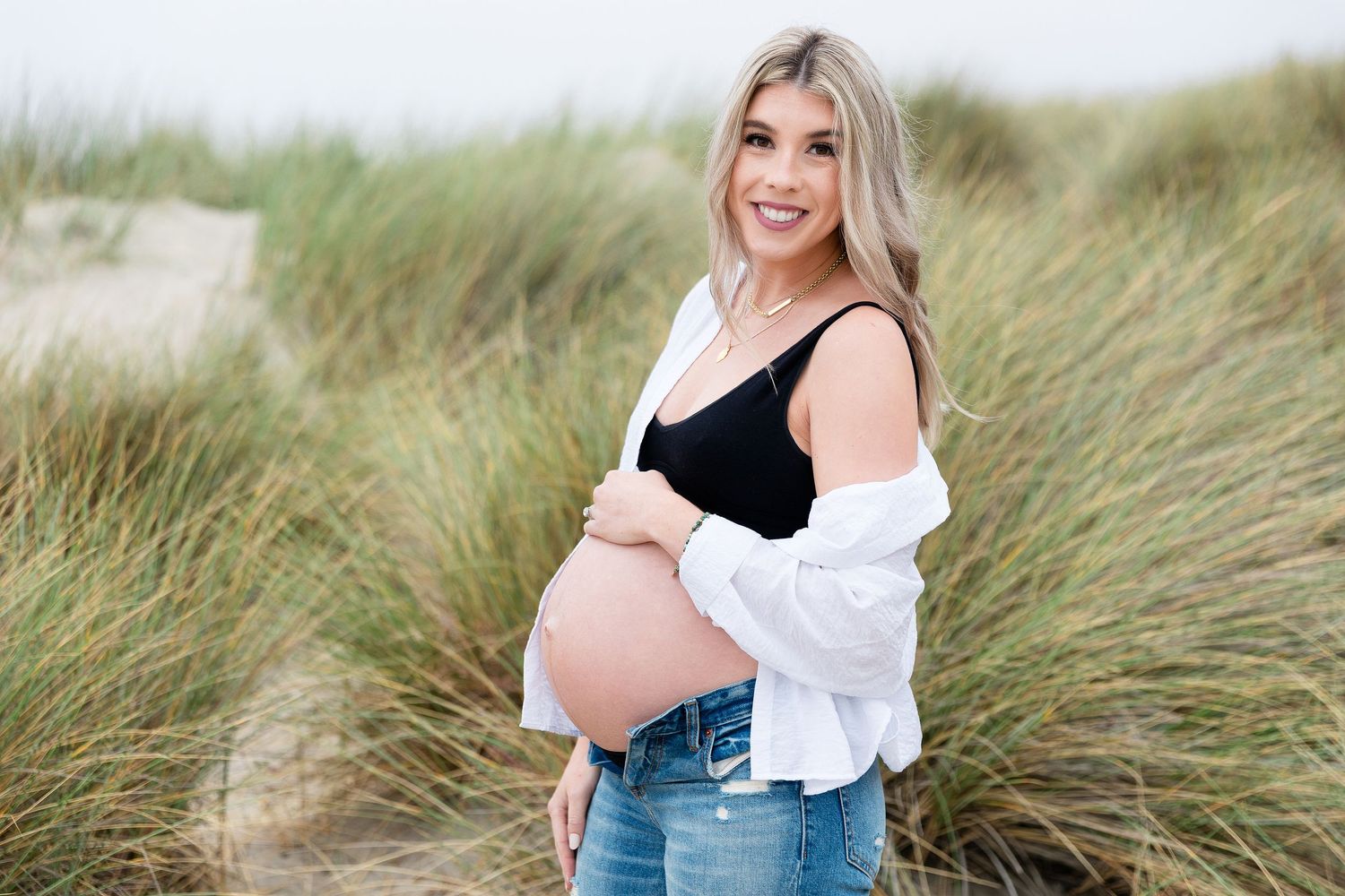 Maternity portrait among beach grass showing woman in casual denim and black top.