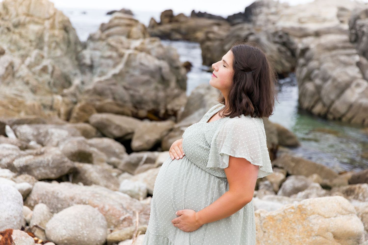 Glowing maternity portrait on rocky beach with waves and coastal backdrop.