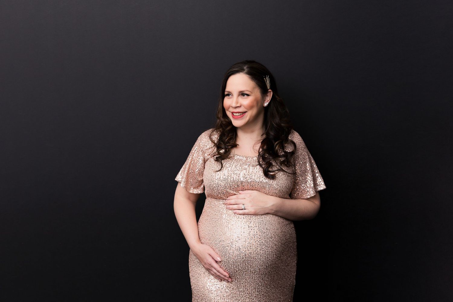Glamorous maternity portrait in shimmering sequined dress against dark backdrop.