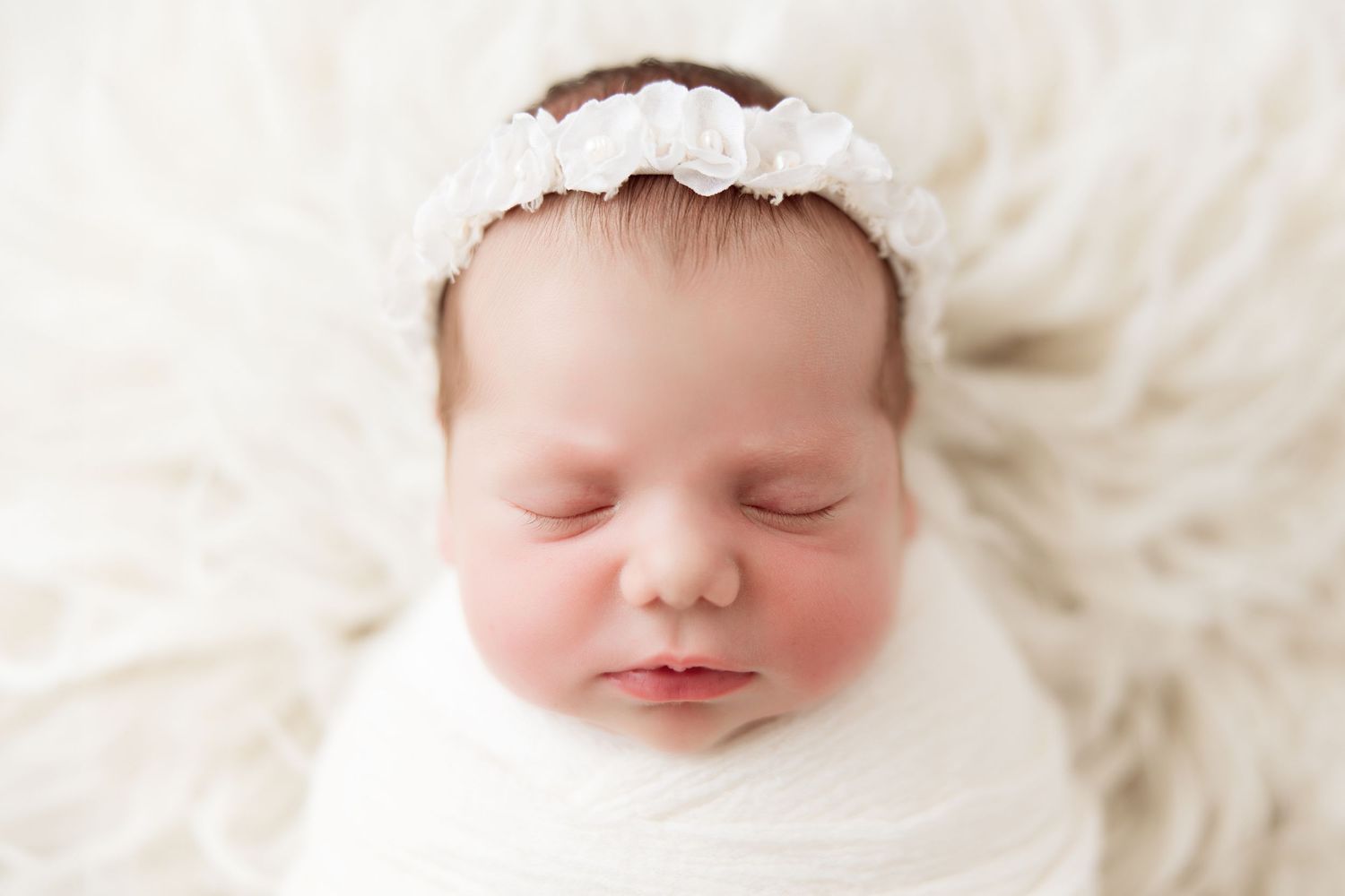 Peaceful sleeping newborn wearing lace headband against white fur backdrop.