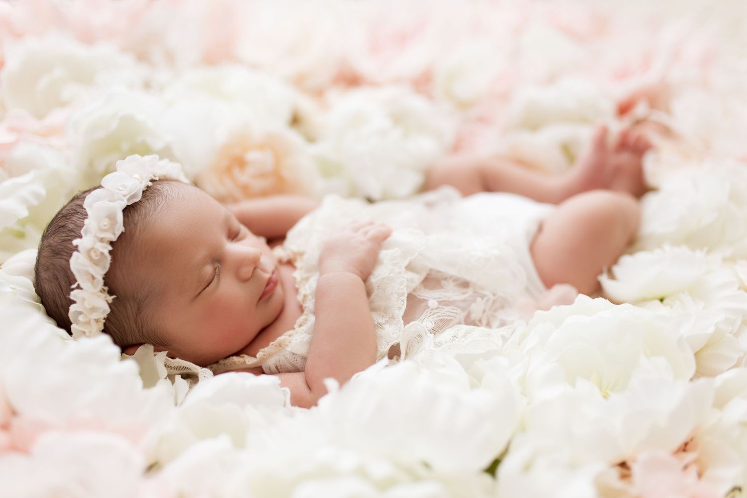 A sleeping newborn baby wearing a white lace outfit rests on soft white and pink floral bedding.