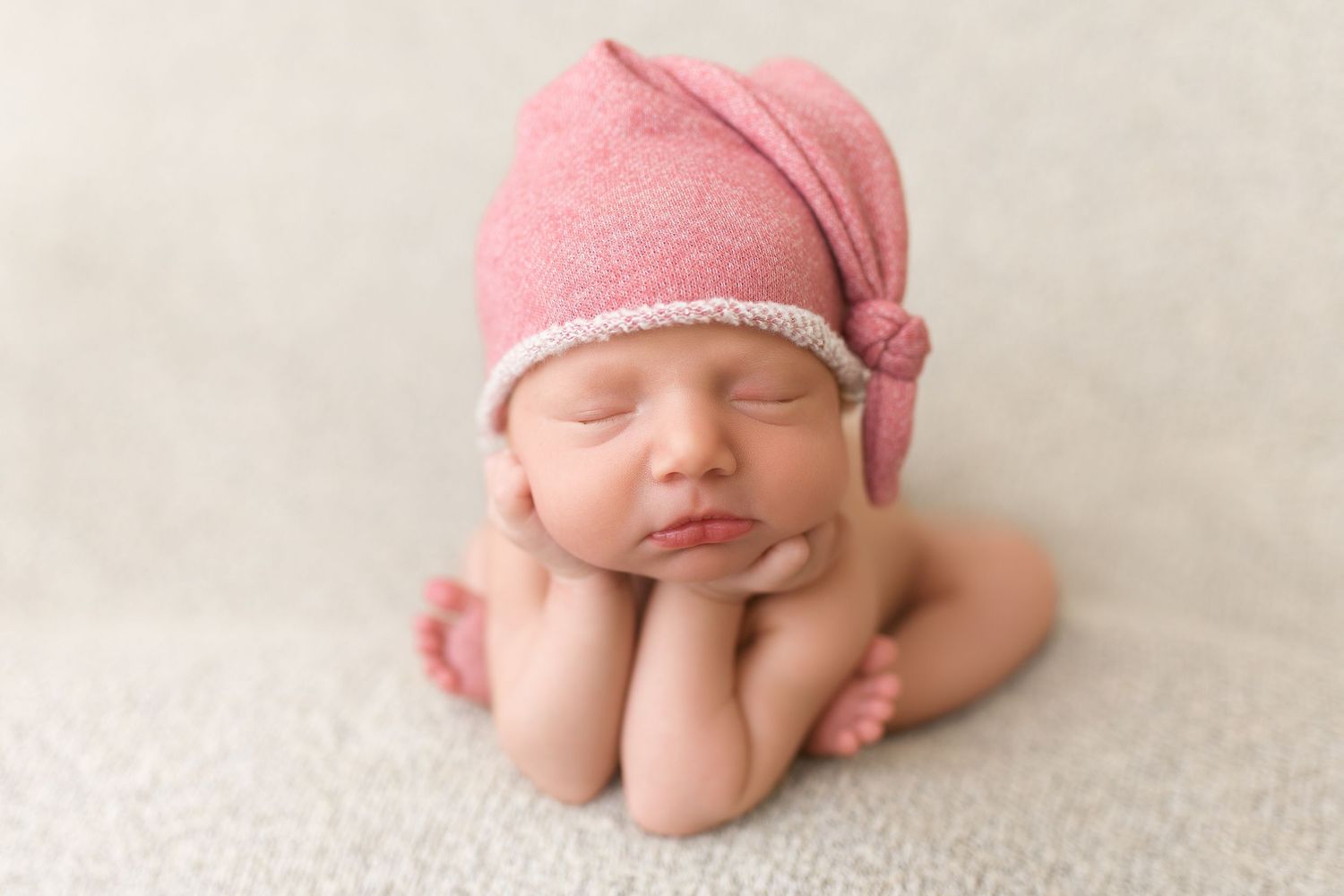 Sleeping newborn wearing pink knit cap posed with hands under chin.