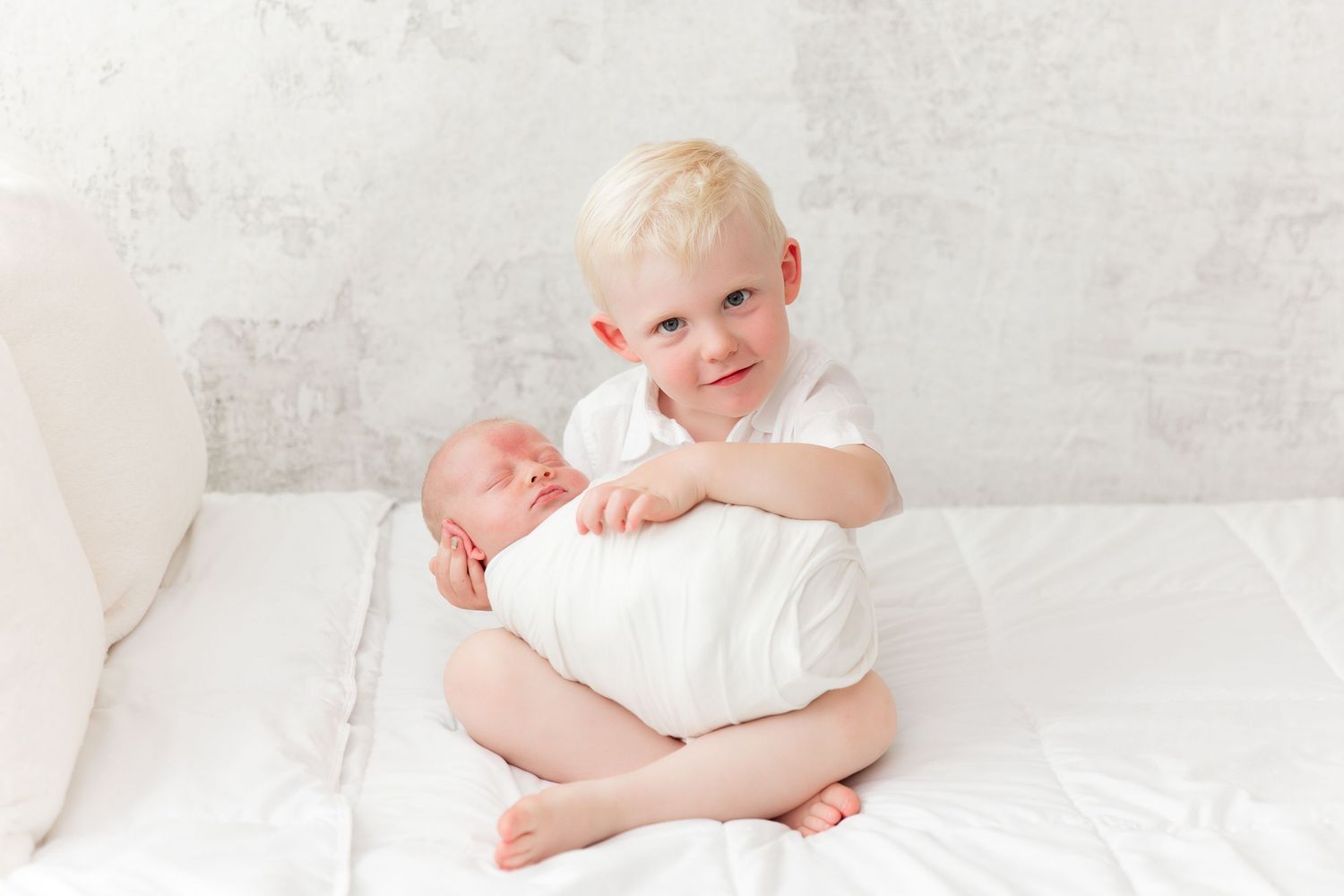 Siblings bonding on white couch with newborn.