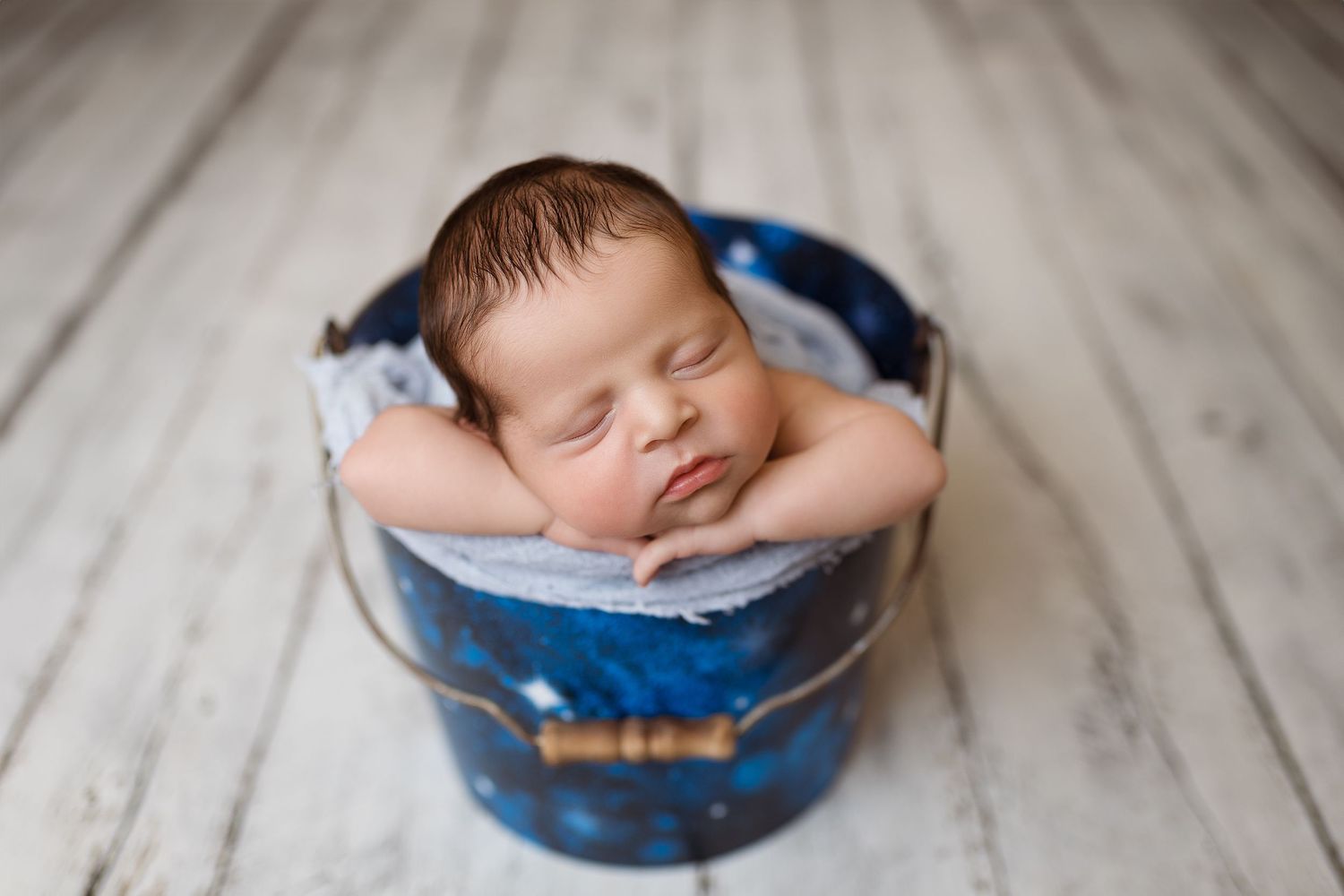 Newborn baby sleeping peacefully in rustic blue bowl on wooden floor.