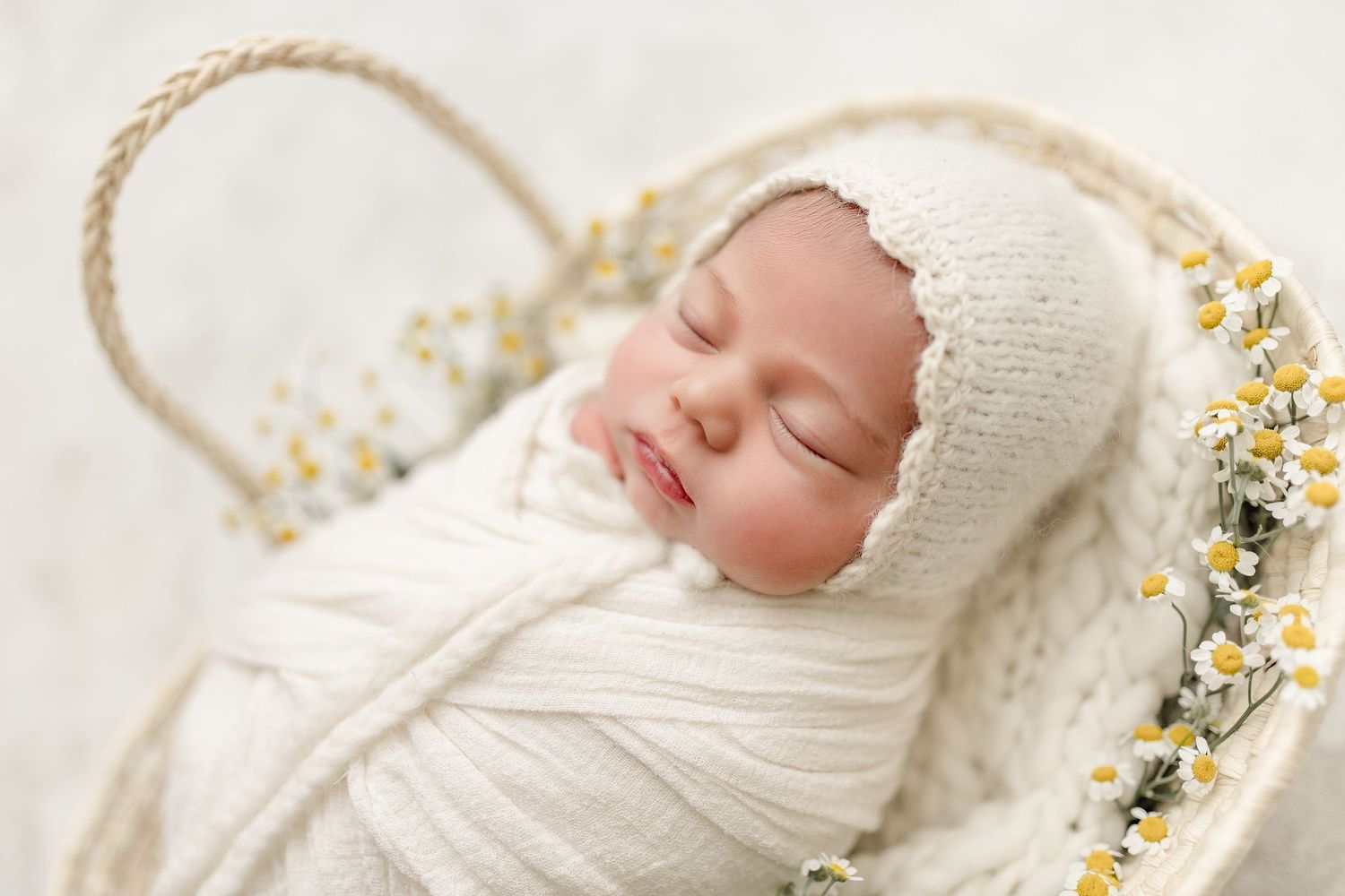 Sleeping newborn wrapped in white swaddle with knit bonnet lying in basket decorated with yellow flowers.
