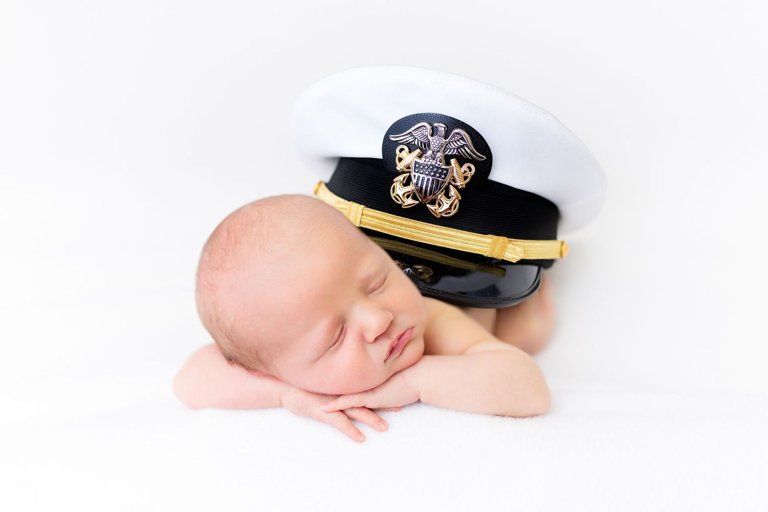 Sleeping baby wearing a white US Navy officer's cap rests peacefully on white background.