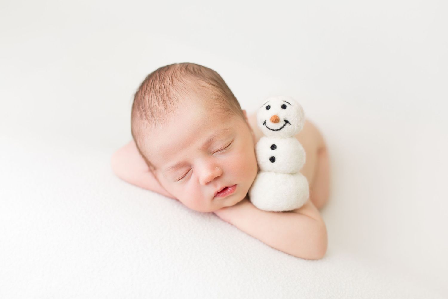 A sleeping newborn rests peacefully next to a small white snowman decoration on a light background.