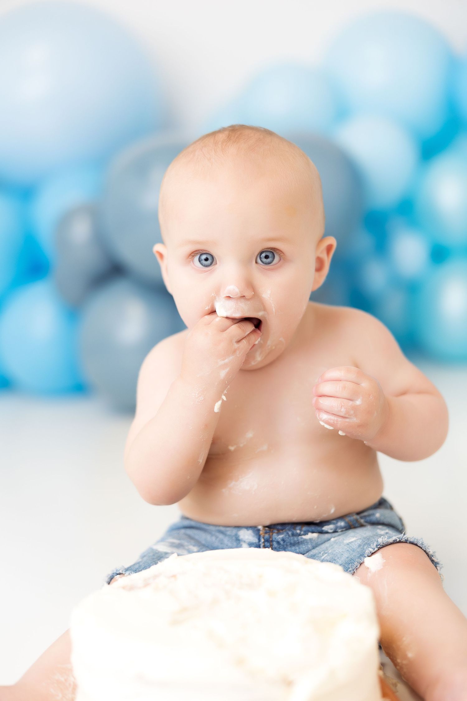 Baby getting messy with white cake during first birthday photoshoot with blue balloon backdrop.