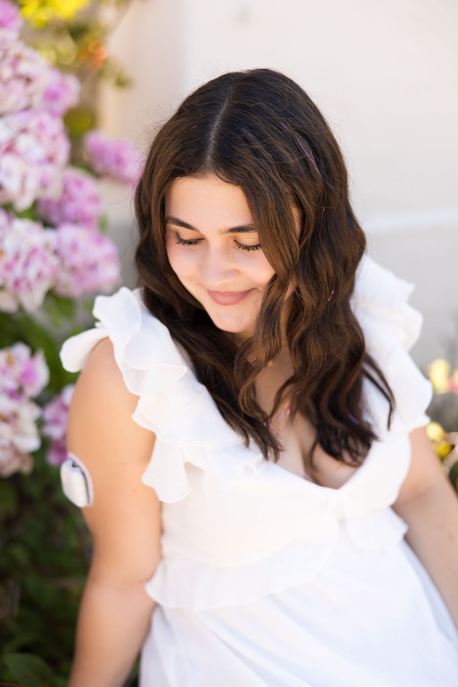 Young woman in white ruffled dress smiling downward surrounded by pink hydrangeas outdoors.