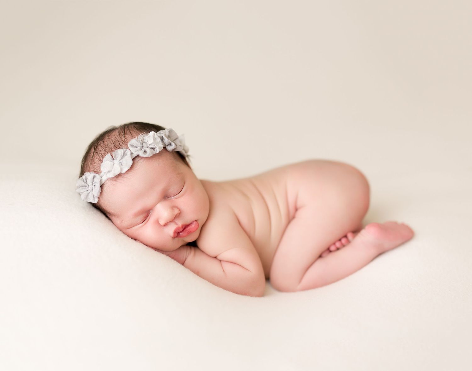 Sleeping newborn with lace headband poses on cream-colored backdrop.