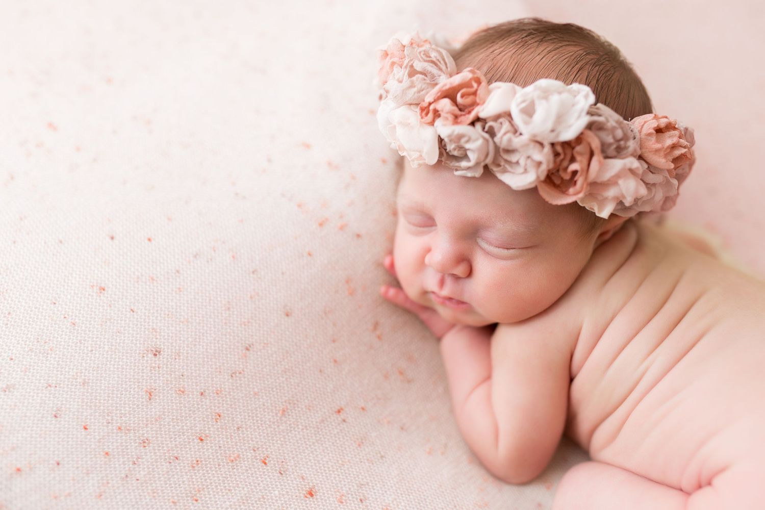 Peaceful newborn wearing floral crown sleeps on pink background with sparkly details.