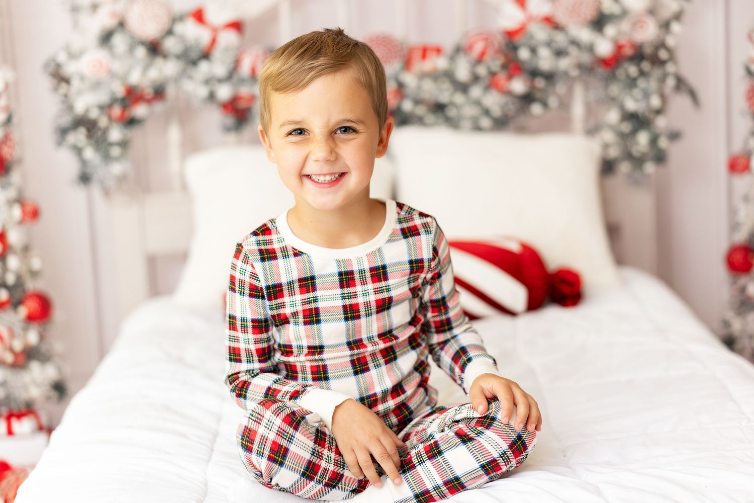 Child in plaid Christmas pajamas sitting cross-legged on white bed with festive decor.