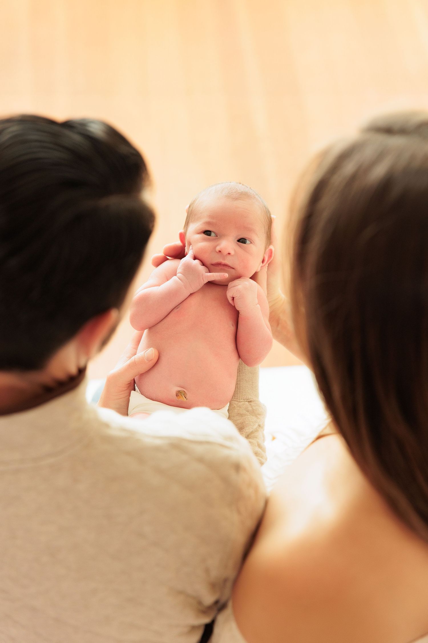 Parents hold their alert newborn baby up between them against warm background.