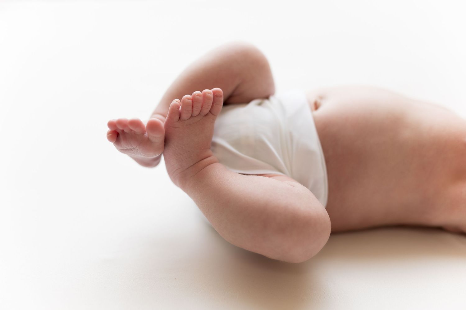 Close up of baby legs and feet in white diaper against light background.