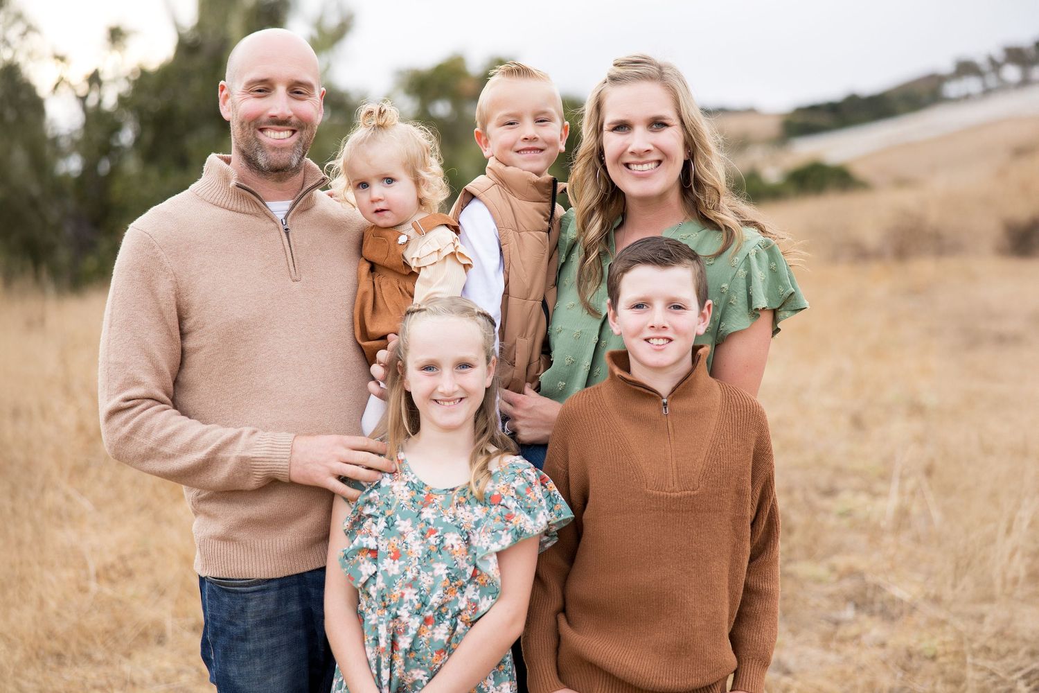 Family members pose together in coordinated earth-tone outfits against a natural outdoor backdrop.