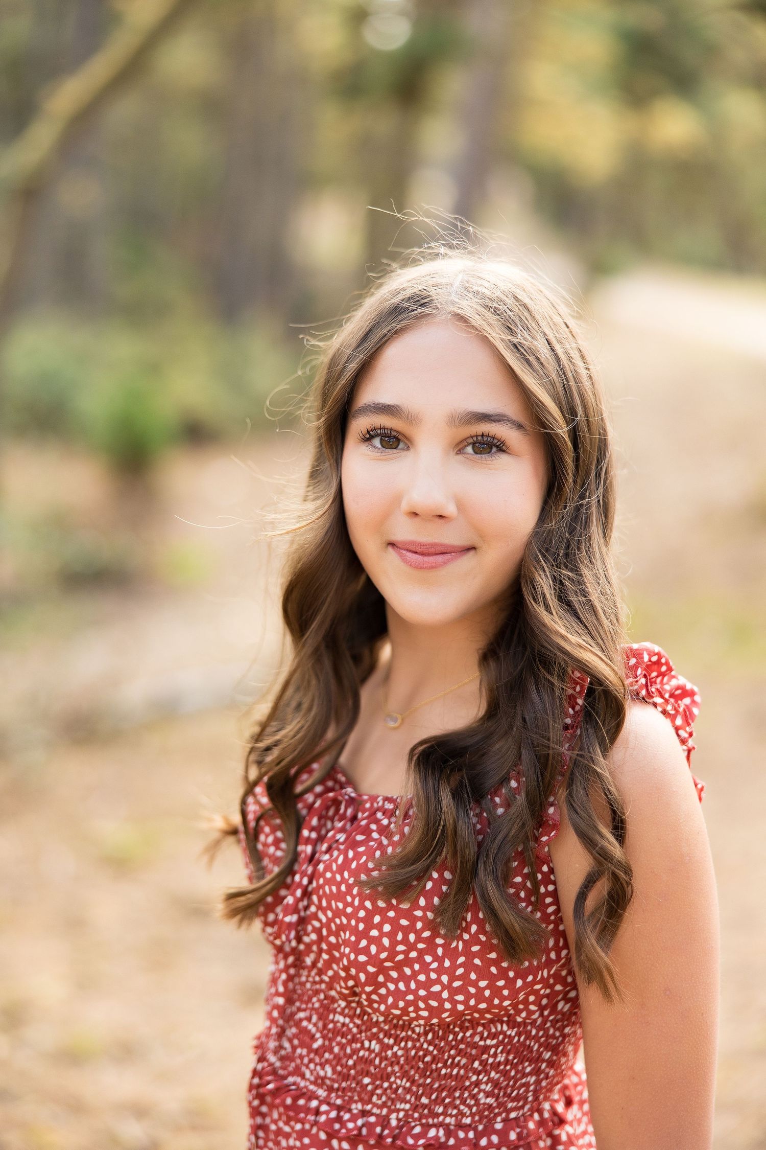 A person in a red floral dress smiles at the camera in a sun-dappled forest setting.