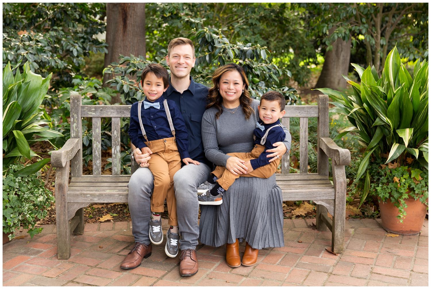 A family sits together on a wooden bench in a lush garden setting with brick pavers and tropical plants.